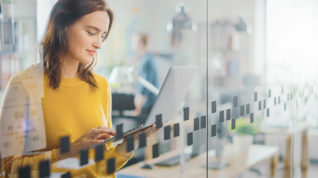photo : Portrait of Beautiful Young Woman with Red Hair Wearing Yellow Sweater Uses Modern Laptop while Standing. Successful Woman Working in Bright Diverse Office.