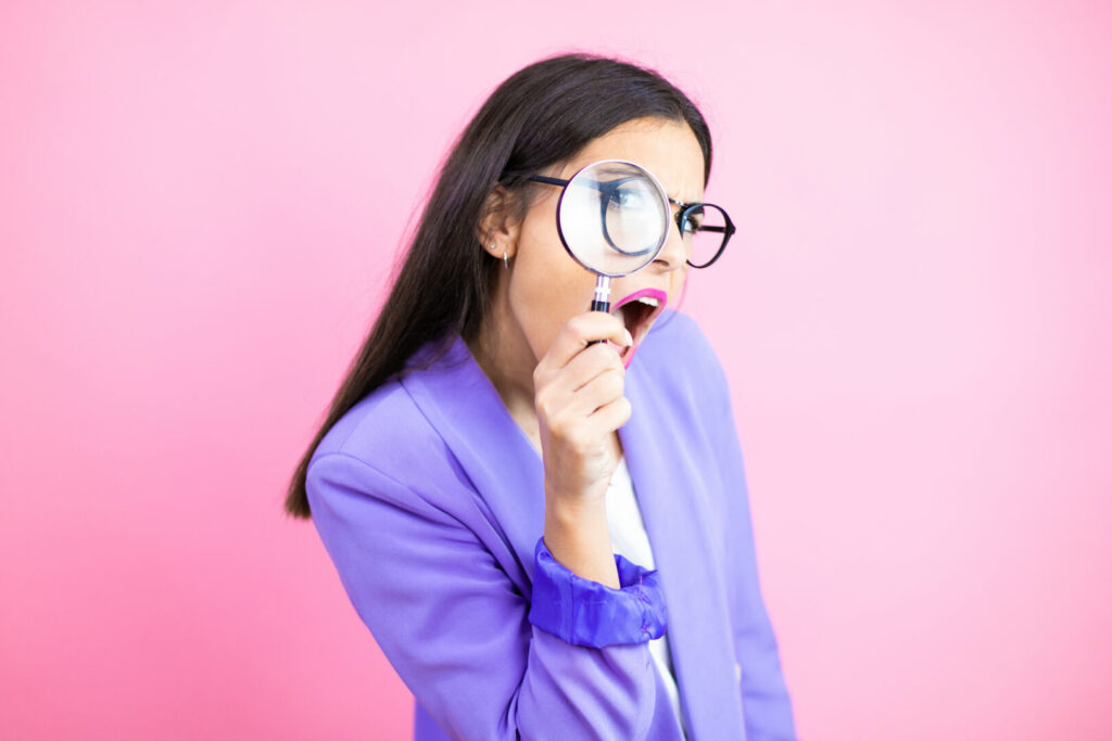 photo : Young business woman wearing purple jacket over pink background surprised looking through a magnifying glass