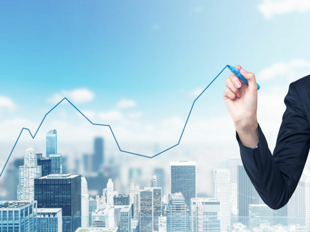 photo : A hand is drawing a growing line graph on the glass screen. New York City space on the background.