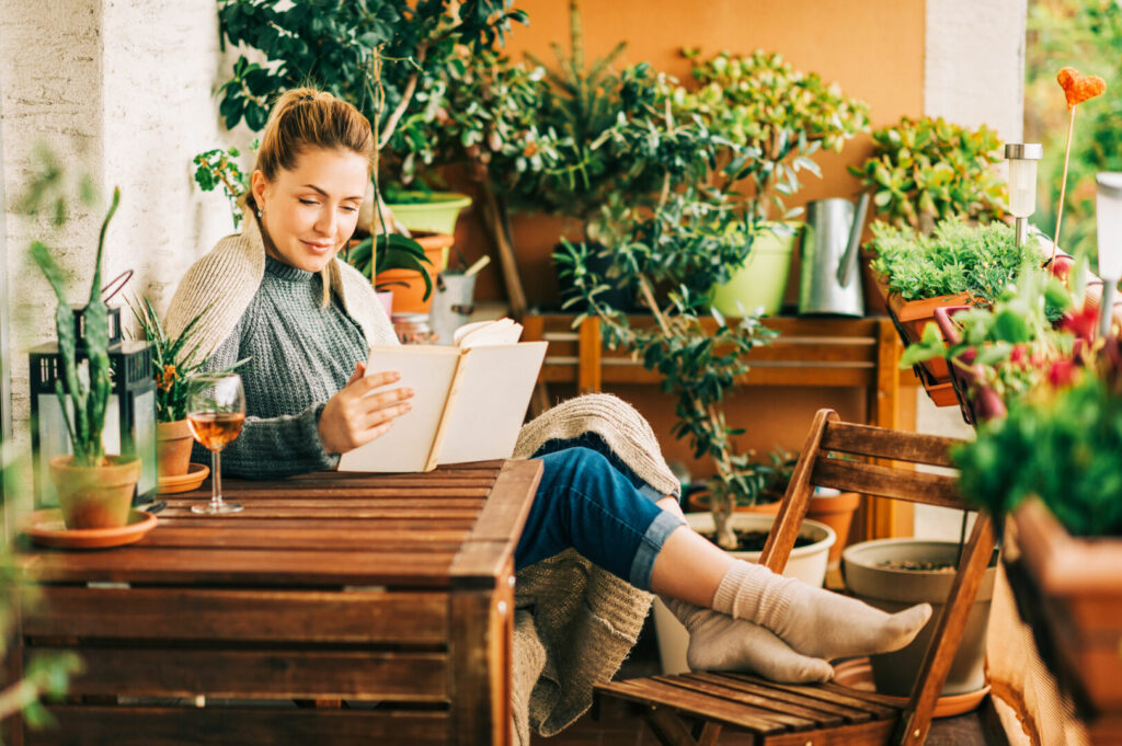 photo : Young beautiful woman relaxing on cozy balcony, reading a book, wearing warm knitted pullover, glass of wine on wooden table